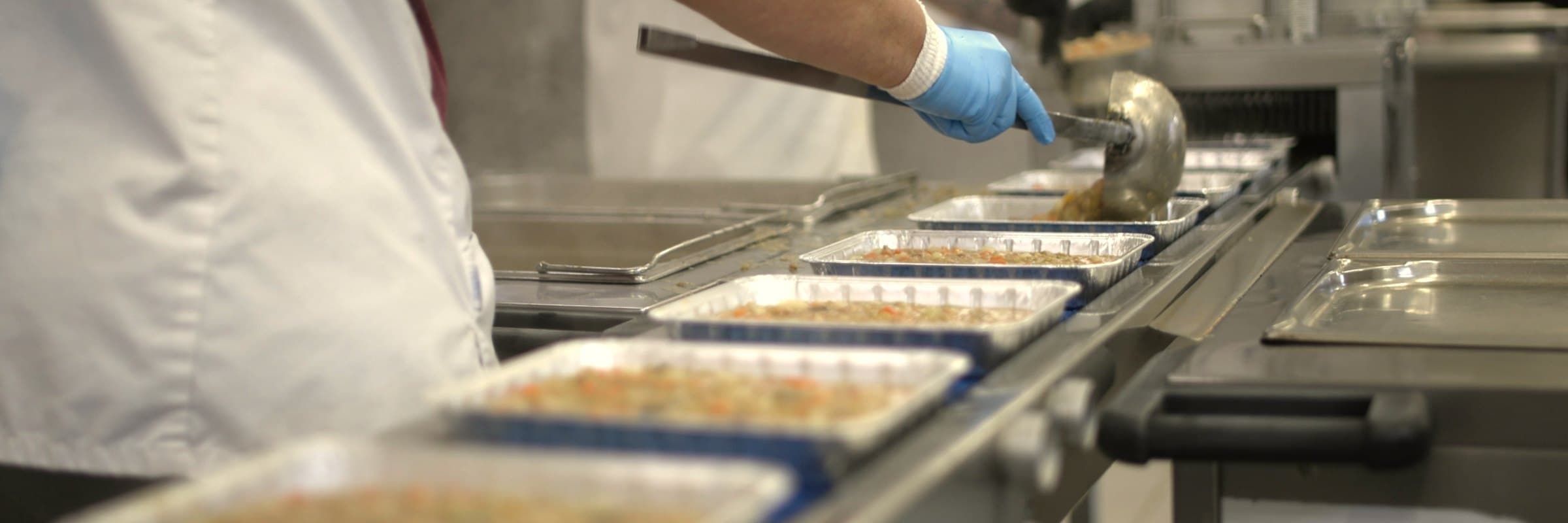 Hands ladling food into trays on a production line in a central kitchen