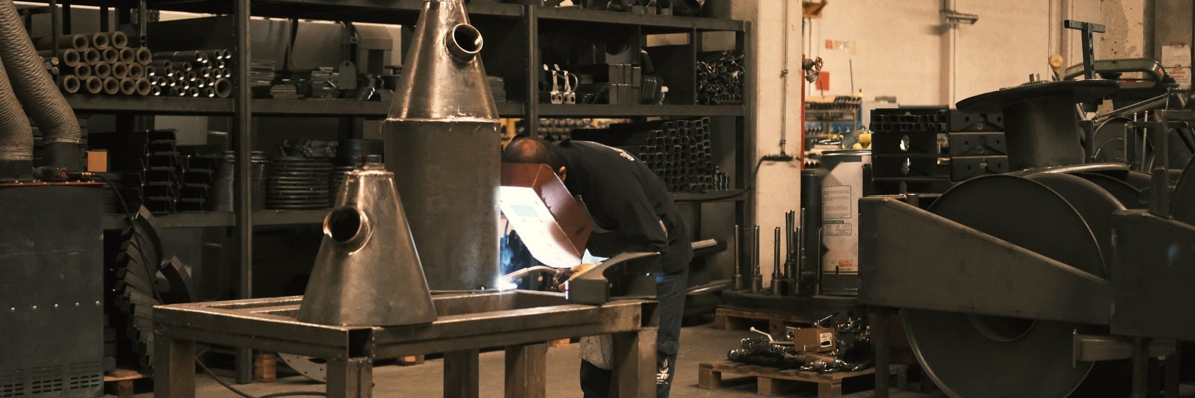 A welder at work in a small metal workshop, among finished parts